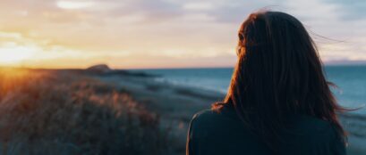 back of a woman standing on a beach and looking at the sunset