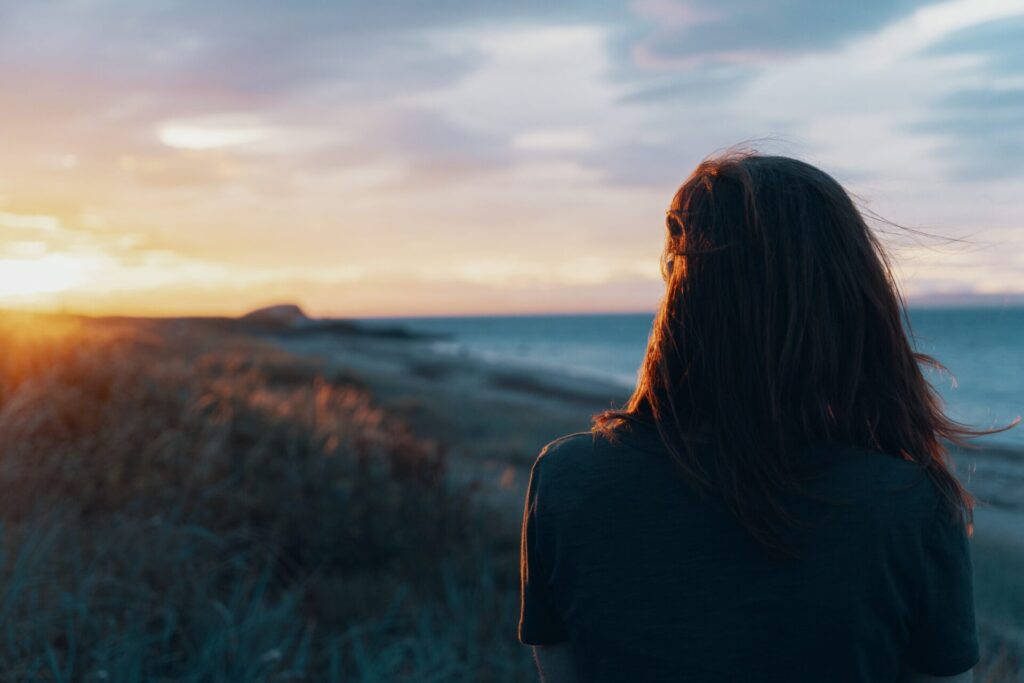 back of a woman standing on a beach and looking at the sunset