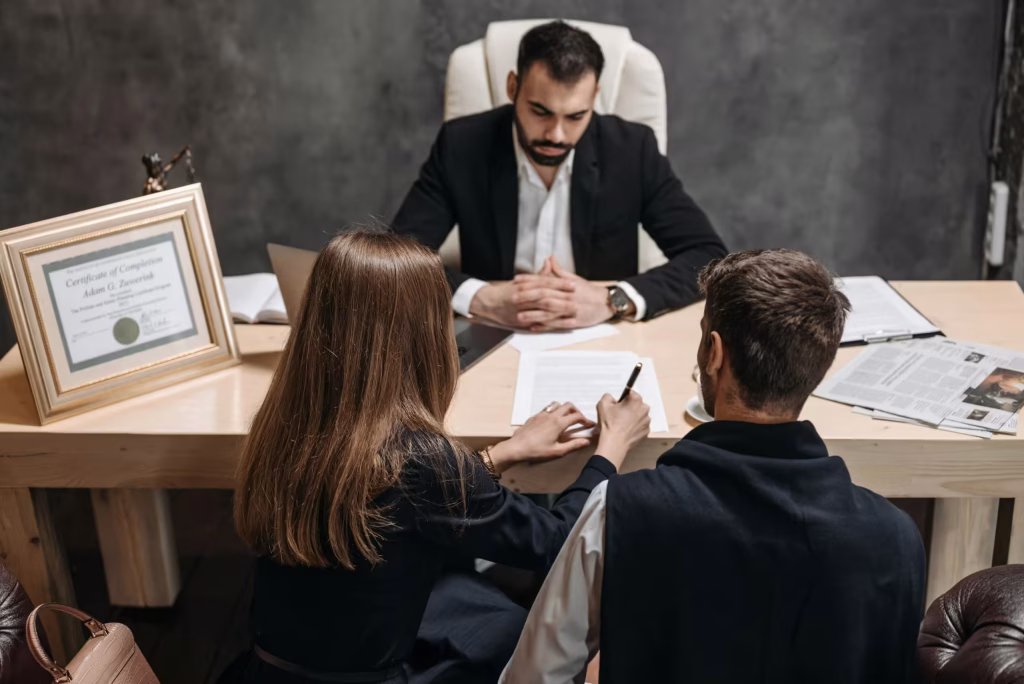 A man and women sitting on a desk and writing on a paper, with a lawyer infront of them.
